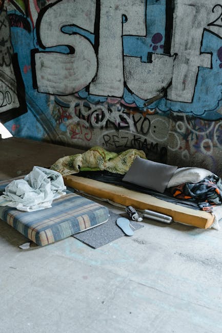 The image depicts a makeshift sleeping area set up on a concrete floor in front of a graffiti-covered wall featuring large, stylized letters in shades of blue, black, and purple. The sleeping arrangement includes a wooden pallet serving as a base, topped with a greenish-yellow blanket and a black jacket or clothing item. Adjacent to the pallet, there is a checked fabric mattress with a small grey foam pad beside it, along with a crumpled white cloth or towel. Near the bedding, a silver laptop is partially covered by clothing, and a white computer mouse and a small cordless electric drill are placed on the ground. The environment appears to be an urban or industrial space often associated with informal or private waste handling, which might be relevant in the context of independent rubbish collection or personal disposal services, as offered by companies such as Rubbish Clearance Maida Vale. The scene emphasizes the need for professional rubbish removal services when managing waste in such environments, particularly for furniture, bedding, and electronic items that are often encountered in private or alternative waste clearance scenarios.