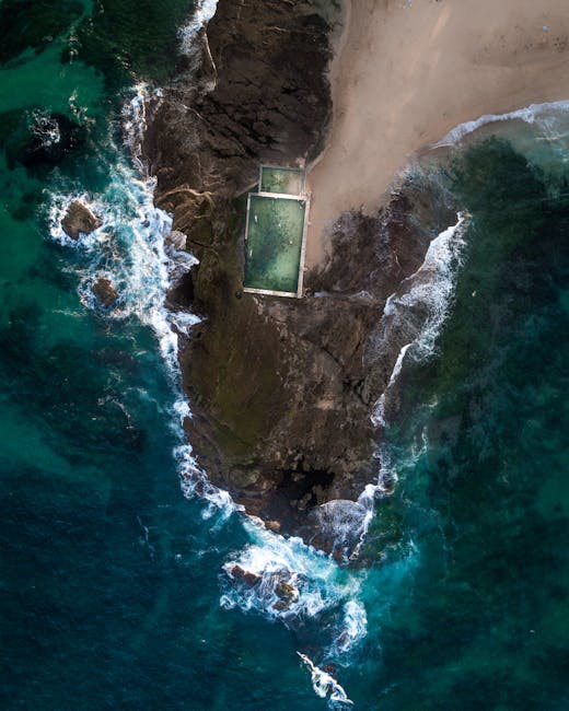 An aerial view of a secluded coastal area showing a small sandy beach adjacent to rocky cliffs, with the ocean waves gently crashing onto the shoreline. The image features a rectangular swimming pool situated on a narrow strip of land between the cliffs and the beach, with clear, greenish water that contrasts against the darker rocks. The surrounding environment includes smooth, weathered rocks, a patch of sandy terrain, and sparse vegetation near the cliffs. The water displays various shades of blue and turquoise, with white foamy surf where the waves break. This scene highlights a quiet, private seaside location, potentially accessible by private or alternative waste handling methods, consistent with the context of independent collection or on-site clearance. The lighting suggests natural daylight, casting subtle shadows on the rocks and highlighting the textures of the shoreline and water surface.