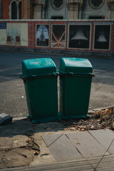 Two green plastic waste bins with black handles and lids are positioned side by side on a paved sidewalk curb in an urban setting, with the left bin slightly taller than the right. The bins have a smooth, glossy surface with a slightly curved top. They are placed on a concrete curb that shows some cracks, dirt, and scattered dry leaves at their base. Behind the bins, there is an asphalt road that appears slightly worn, with a faint white line marking the edge of the pavement. In the background, a brick and plaster building with arched windows and decorative architectural elements is visible, along with a construction fence or billboard featuring partially obscured posters or artwork. The scene suggests an exterior location where waste disposal options are accessible apart from municipal rubbish collection, aligning naturally with private or independent waste handling services such as those provided by Rubbish Clearance Maida Vale.