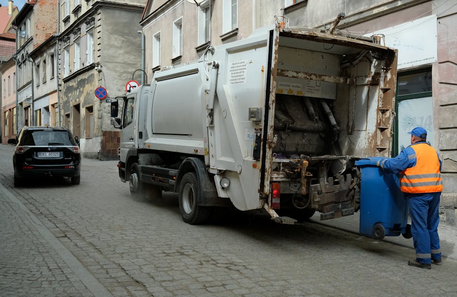 A worker dressed in a high-visibility orange vest and blue uniform with a matching blue cap is seen operating a blue wheeled trash bin, positioning it towards the rear of a white waste collection truck parked on a narrow urban street. The truck has a large, enclosed waste compartment made of metal with visible signs of rust and use, including a partially open rear hatch revealing the crushing and compacting mechanisms inside. The street is paved with cobblestones and lined with older, multi-story buildings with weathered exteriors, some showing patches of peeling paint and minor repairs. To the left, a black car is parked along the curb, and there are traffic signs mounted on a pole indicating parking restrictions. The scene is lit by natural daylight, with no shadows indicating an overcast or diffuse light environment. This setup reflects an on-site waste collection process, often undertaken as an alternative or supplement to regular rubbish removal services offered by companies like Rubbish Clearance Maida Vale, supporting private disposal or on-demand rubbish clearance for urban settings.