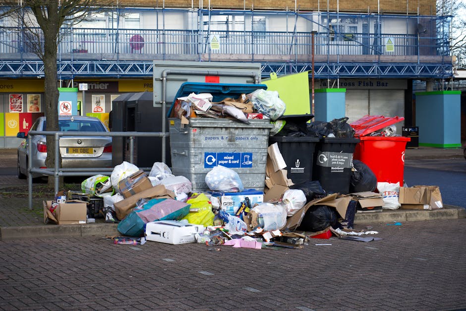 The image depicts a collection of overflowing rubbish bins and scattered waste on a paved area near a parking lot. The central focus is on a large grey mixed paper and card bin, which is open and filled with crumpled paper, cardboard, and packaging materials, with some waste spilled onto the ground around it. To its right, there are black recycling bags and a bright red refuse bin, both overflowing with assorted waste including plastic and paper materials. In front of the bins, various disposed items such as cardboard boxes, plastic bags, and packaging material are spread across the pavement, contributing to an untidy appearance. On the left side, a silver car is parked close to a green metal railing that separates the waste area from the pedestrian walkway. Behind the waste collection point, a building under construction or renovation is visible, featuring a blue metal scaffolding structure and windows, with some signage indicating commercial premises. The environmental context suggests an urban or suburban setting where private waste collection might take place, supported by the presence of multiple waste containers and scattered rubbish. The scene is captured during daylight hours, with natural lighting highlighting the textures and clutter of the discarded waste.