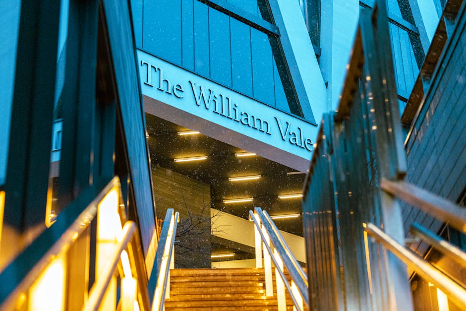 A view looking up at the entrance to The William Vale, a modern hotel building with a glass and metal facade, illuminated by warm yellow lighting. The structure features angular architectural elements with large glass panels reflecting the surrounding environment. Prominent signage displaying the hotel name is situated near the top of the entrance, partially visible in the image. The overall scene is set during nighttime, with the exterior lights creating a contrast against the dark sky. The vantage point emphasizes the building’s contemporary design, which may relate to private property or commercial premises that could require proactive rubbish or waste management services provided by companies like Rubbish Clearance Maida Vale for maintaining clean and orderly surroundings.
