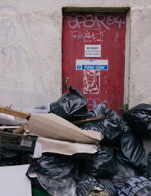 The image depicts a collection of overflowing rubbish bins and scattered waste on a paved area near a parking lot. The central focus is on a large grey mixed paper and card bin, which is open and filled with crumpled paper, cardboard, and packaging materials, with some waste spilled onto the ground around it. To its right, there are black recycling bags and a bright red refuse bin, both overflowing with assorted waste including plastic and paper materials. In front of the bins, various disposed items such as cardboard boxes, plastic bags, and packaging material are spread across the pavement, contributing to an untidy appearance. On the left side, a silver car is parked close to a green metal railing that separates the waste area from the pedestrian walkway. Behind the waste collection point, a building under construction or renovation is visible, featuring a blue metal scaffolding structure and windows, with some signage indicating commercial premises. The environmental context suggests an urban or suburban setting where private waste collection might take place, supported by the presence of multiple waste containers and scattered rubbish. The scene is captured during daylight hours, with natural lighting highlighting the textures and clutter of the discarded waste.