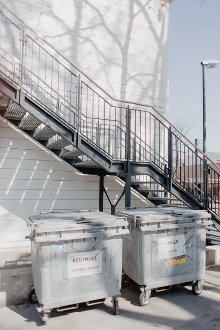 A pair of large, grey wheeled waste bins positioned on a paved outdoor surface in front of a white wall. The bins are made of hard plastic with textured surfaces and are labeled with different waste types, one indicating 'RESIDUAL WASTE' and the other for paper recycling. They sit beneath an external metal staircase with a black railing, which extends upward along a building facade. The staircase features metal steps with a grated surface and has shadow patterns cast by nearby trees on the wall behind. To the right, a tall street lamp is visible against a clear, blue sky. The scene suggests an urban setting possibly used for communal or commercial waste collection, with the presence of outdoor waste bins implying a private collection service that could complement or serve as an alternative to municipal rubbish removal for flat or building waste disposal needs, as seen in typical Maida Vale properties managed by rubbish clearance services like Rubbish Clearance Maida Vale.