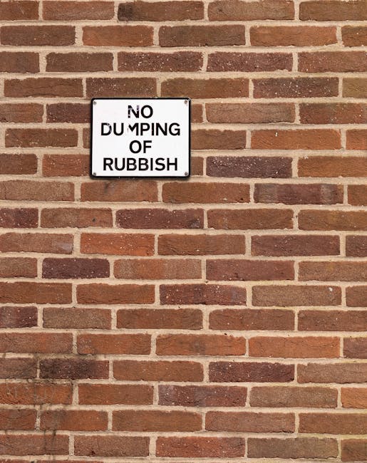 Close-up of a white rectangular sign mounted on a red-brown brick wall, with black text that reads 'NO DUMPING OF RUBBISH' in uppercase letters, with the words slightly offset and unevenly spaced. The brick wall has a rough texture with visible mortar lines and varies in shade from light to dark red and brown. The sign is centrally positioned in the upper half of the image, emphasizing the message against the background of the brick surface. The scene appears to be outdoors, possibly on the side of a building, highlighting the importance of private rubbish management and preventing illegal dumping in areas like Maida Vale. The lighting is neutral with minimal shadows, providing a clear view of both the sign and the brick wall's detail, supporting themes of waste regulation and vigilant rubbish clearance services by companies such as Rubbish Clearance Maida Vale.