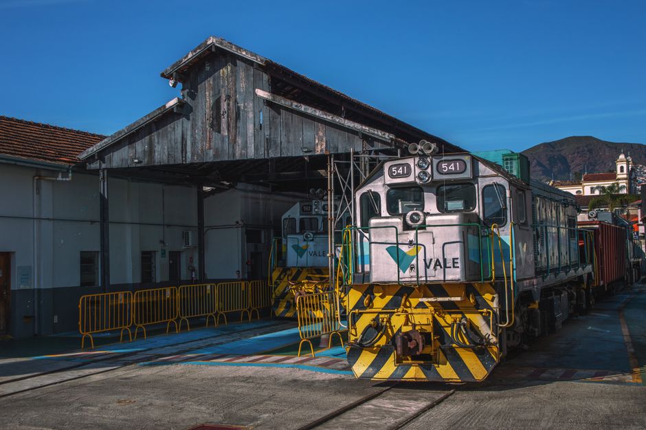A silver diesel locomotive with the number 541 painted on its front is positioned on railway tracks in front of a weathered wooden shed with a corrugated metal roof, situated outdoors on a clear day. The train features yellow and black diagonal safety stripes on its front lower section, and the logo of Vale is visible on its front and side. To the left, a small white building with windows and a door can be seen, partially obscured by the train. Surrounding the train, there are orange safety barriers along the ground, and the environment includes distant mountains and a partly cloudy sky, indicating a rural or industrial setting suitable for private waste or materials handling, relevant to rubbish removal services such as those provided by Rubbish Clearance Maida Vale.