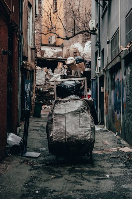 An urban alleyway cluttered with a large, wheeled rubbish sack in the foreground, made of grey, woven plastic material with visible seams and creases. The alley is flanked by brick and metal-clad buildings on either side, with graffiti marking the walls on the right. In the background, a pile of mixed waste materials, including cardboard boxes, plastic bags, and other debris, is stacked against a raised concrete edge or wall, partially obscured by leafless tree branches overhead. The uneven, dark asphalt surface of the alley shows scattered dirt and small debris, with some areas darker and damp. The scene is lit by natural daylight, giving a muted, neutral tone that highlights the disorder typical of an independent rubbish collection site, often associated with private waste handling services like those offered by Rubbish Clearance Maida Vale. The overall visual suggests the alley is used for temporary waste storage or clearance, emphasizing the need for professional rubbish removal services to manage such clutter efficiently.