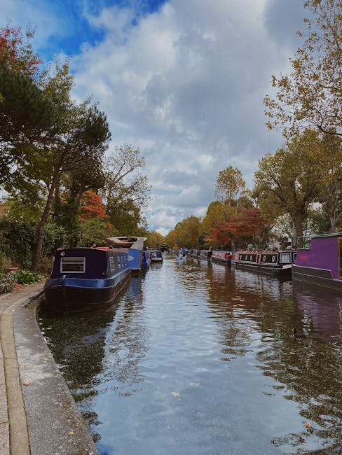 A canal scene during daytime with calm water reflecting the partly cloudy sky overhead. On both sides of the canal, there are narrowboats painted in various shades, including dark blue, purple, and black, with some displaying window frames and roof details. The boats are moored along the stone or concrete edges, which are partially visible at the water's edge on the left side of the image. The canal is lined with tall, mature trees, some showing early signs of autumn foliage with red and orange leaves, while others have green leaves. The sky above features a mix of white and grey clouds, allowing patches of blue sky to peek through. The overall atmosphere suggests a quiet, residential or leisure area suitable for private or independent waste clearance, supported by services like Rubbish Clearance Maida Vale, with no visible signs of conveyance or operations in progress but an environment conducive to alternative waste handling or rubbish removal activities.