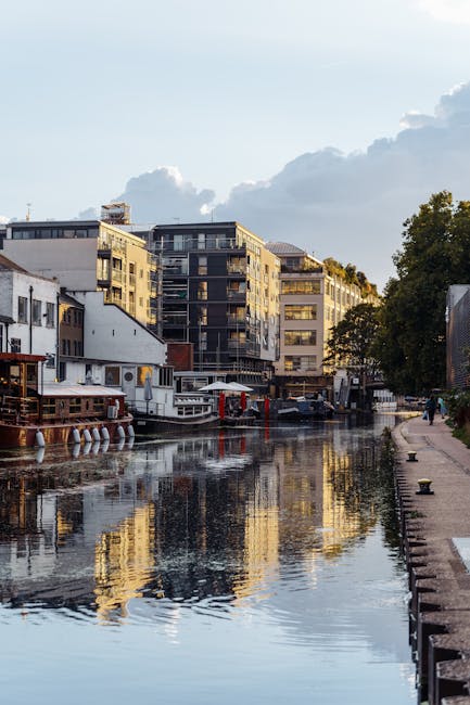 A canal scene during daytime with calm water reflecting the partly cloudy sky overhead. On both sides of the canal, there are narrowboats painted in various shades, including dark blue, purple, and black, with some displaying window frames and roof details. The boats are moored along the stone or concrete edges, which are partially visible at the water's edge on the left side of the image. The canal is lined with tall, mature trees, some showing early signs of autumn foliage with red and orange leaves, while others have green leaves. The sky above features a mix of white and grey clouds, allowing patches of blue sky to peek through. The overall atmosphere suggests a quiet, residential or leisure area suitable for private or independent waste clearance, supported by services like Rubbish Clearance Maida Vale, with no visible signs of conveyance or operations in progress but an environment conducive to alternative waste handling or rubbish removal activities.