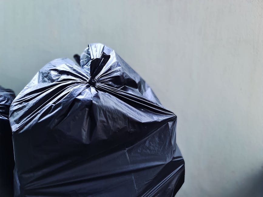 Close-up of three black plastic rubbish bags, tightly knotted at the top, with glossy, slightly crinkled surfaces reflecting light. The bags are stacked against a plain, light-colored wall in an outdoor or semi-enclosed environment, possibly a driveway or alleyway. The background is simple and uncluttered, emphasizing the bags' material and texture. The photograph captures the bags in a clean, organized manner, typical of private waste disposal or rubbish removal services, such as those offered by Rubbish Clearance Maida Vale. The overall scene conveys a straightforward example of waste collection for a domestic or commercial property, with no additional objects or environmental details visible in the frame. The natural lighting highlights the bags' shiny finish and creases, suggesting daylight or ambient outdoor illumination. This image is suitable for illustrating on-site rubbish clearance or private waste management practices, aligning with the context of alternative rubbish collection methods in urban settings.