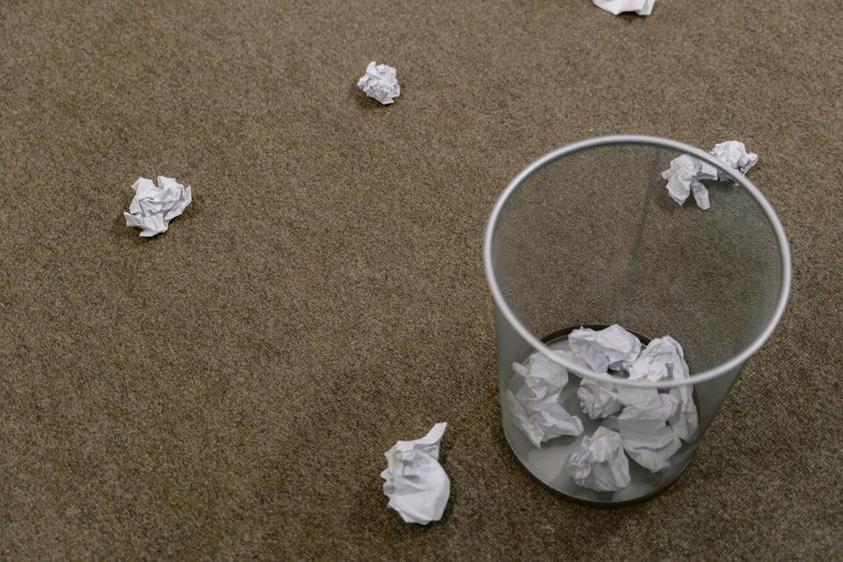 A clear cylindrical waste bin made of transparent plastic sits on a brown, textured carpeted floor, partially filled with crumpled white paper. Several crumpled pieces of white paper are scattered around the bin on the floor, with some near the rim of the bin and others further away. The scene is illuminated by natural or diffused lighting, creating soft shadows and highlights on the paper and the bin. The setting appears to be an indoor office or domestic space, illustrating common waste disposal in a private environment. The image relates to rubbish removal services, highlighting the process of individual waste collection and the importance of maintaining clean and tidy work or living areas, a core aspect of private on-site clearance handled by companies like Rubbish Clearance Maida Vale.