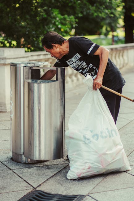 A middle-aged man with dark hair wearing a black T-shirt with white and blue text and graphics, leaning forward as he disposes of waste into a metal public litter bin on a paved sidewalk. He is holding a large white plastic bag filled with rubbish with his left hand, while his right hand is extended into the open top of the bin. The bin is composed of stainless steel with a cylindrical shape, featuring a foot pedal on the right side for opening the lid. In the background, there are lush green trees and bushes, casting dappled sunlight on the scene, suggesting a park or public outdoor space. The pavement beneath the man's feet is made up of square concrete slabs, and there is a faint shadow pattern indicating a bright day. This scene exemplifies on-site waste disposal in a public environment, aligning with practices for litter control and the importance of timely rubbish removal, relevant to private waste handling services such as those provided by Rubbish Clearance Maida Vale.