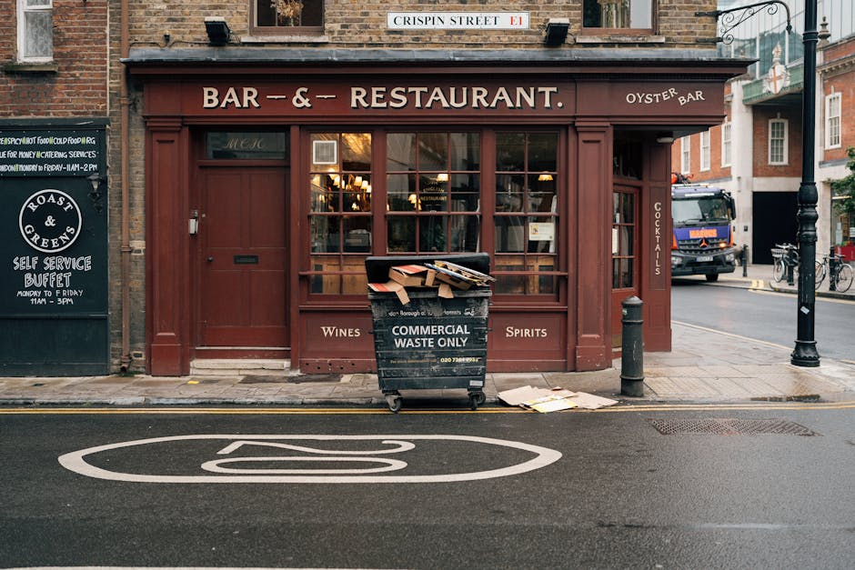 A black commercial waste bin labeled 'Commercial Waste Only' is positioned on the sidewalk in front of a red-brick building with large glass windows, which appears to be a restaurant or bar. The bin contains scattered cardboard boxes and paper debris, with some pieces lying on the ground nearby. The building has a sign indicating it is a bar and restaurant, with the words 'Wines,' 'Spirits,' and 'Cocktails' visible on the facade. To the left of the entrance, there is a blackboard-style sign advertising a self-service buffet and coffee shop operating hours. A bollard is installed on the right side of the bin to prevent vehicle access to the pavement. The street surface in the foreground shows a white painted bicycle lane symbol, and a parked vehicle, including a truck, can be seen in the background on the opposite side of the road. The environment appears urban and busy, typical of a city street in Maida Vale, with street lamps and nearby bicycles, highlighting the context of municipal waste disposal or private clearance services in a commercial area.